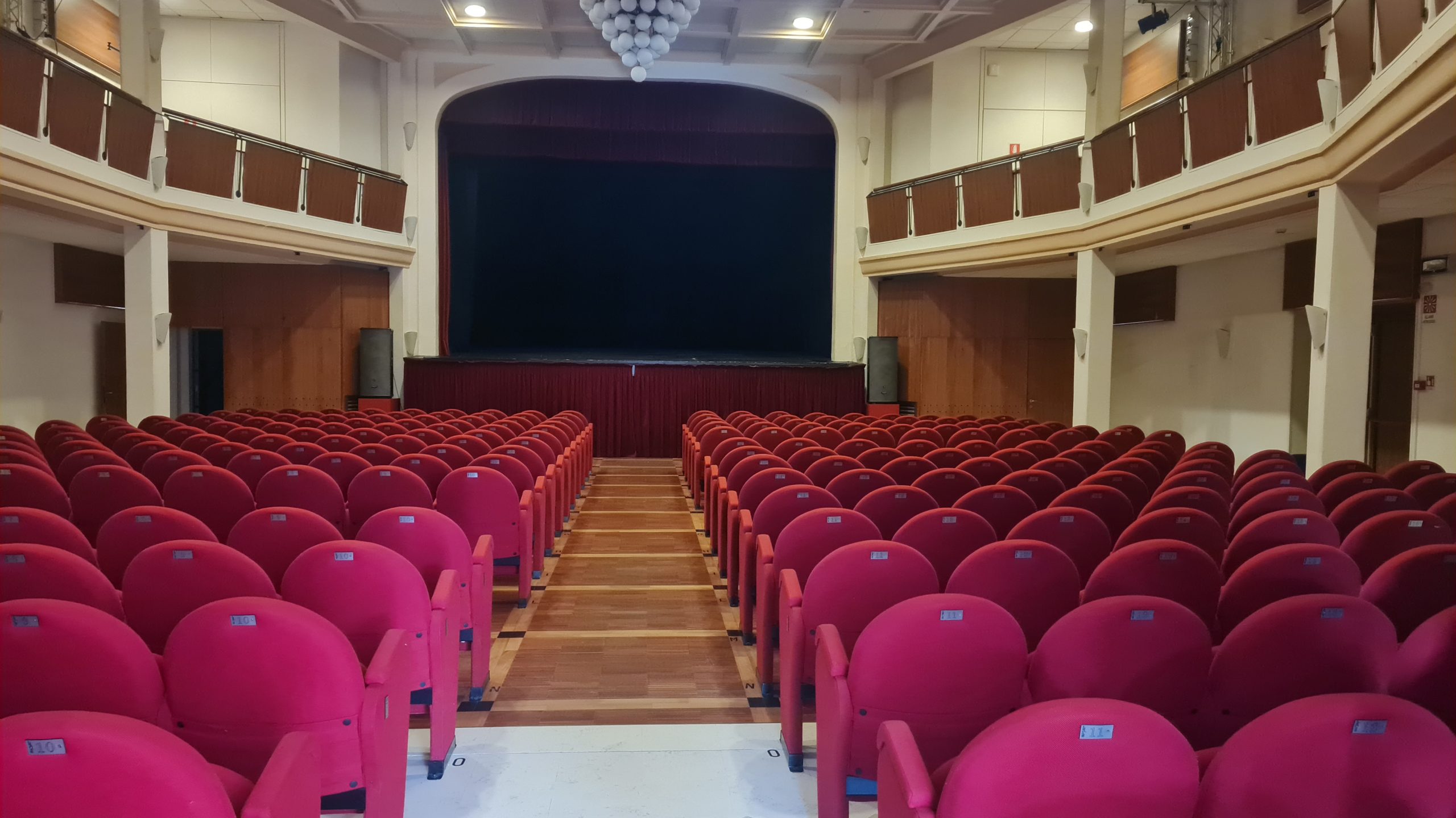 Interno di un teatro con poltrone rosse e palco centrale, vista dalla platea.